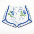 Shorts Verzura III DELAMIA — foglie verdi, fiori blu e bande geometriche laterali su bianco, vista frontale