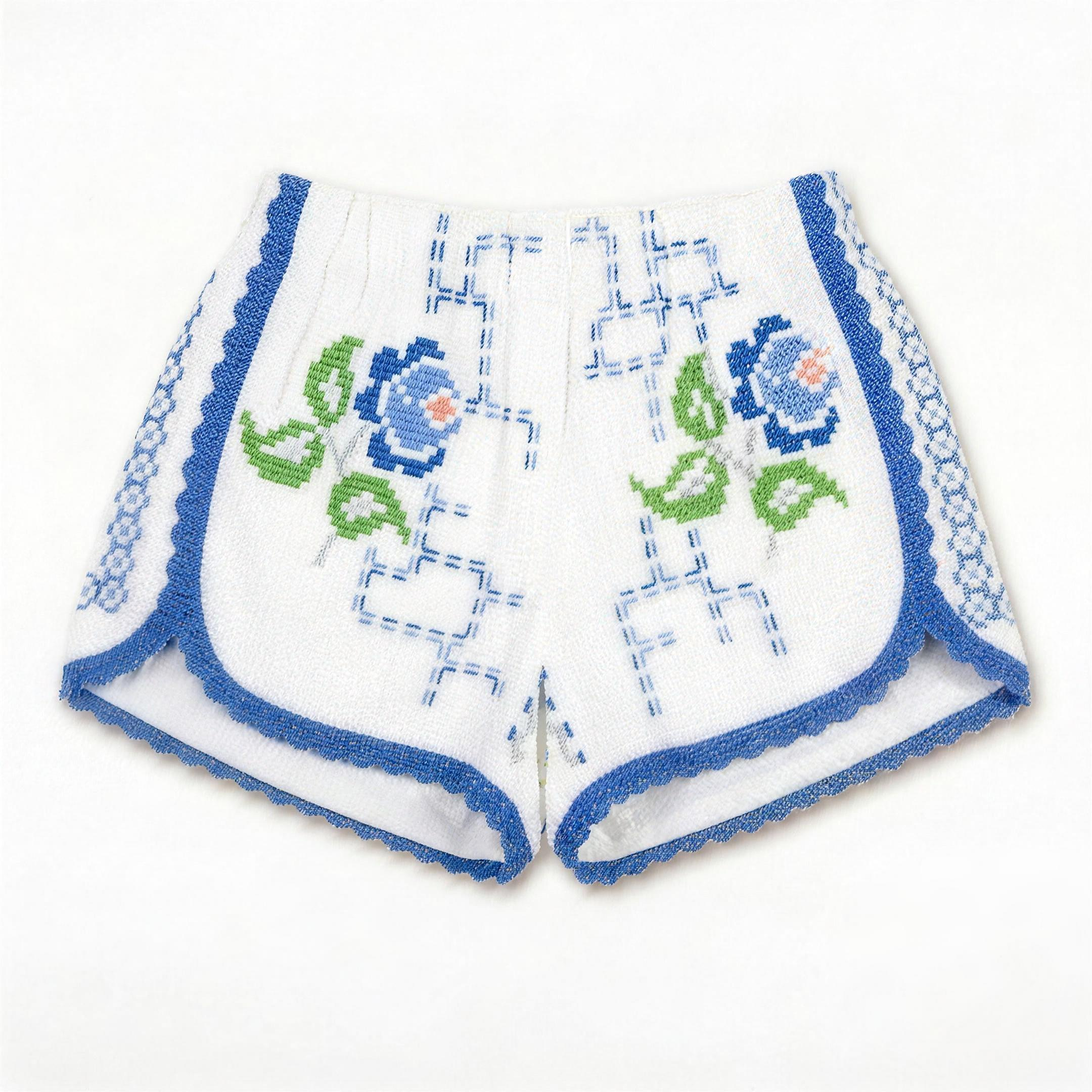Shorts Verzura III DELAMIA — foglie verdi, fiori blu e bande geometriche laterali su bianco, vista frontale
